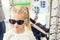 Cute little young caucasian blond girl trying on and choosing sunglasses in front of mirror at optic eyewear store. Adorable