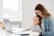 Cute mother hugs baby works from home, she sits in the kitchen and using laptop for remote work, her cute little girl