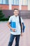 Cute, nice, young 11 years old boy in blue shirt stands with workbooks and backpack in front of his school. Education