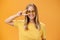 Cute optimistic and friendly young caucasian woman with fair hair in yellow t-shirt and sunglasses showing peace sign