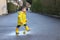 Cute playful little baby boy in bright yellow raincoat and rubber boots playing with toy boat and rubber ducks in small puddle at