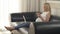 A cute pregnant blonde-haired woman is sitting on the sofa with a laptop and a phone in her hands. She is looking
