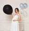 A cute pregnant young woman in anticipation of a baby in a white dress, hugs belly, isolated white neoclassical wall.