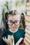 Cute preschool age girl with dimples on her cheeks in round big glasses looking at a camera between two books piles