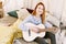 Cute red-haired woman learns to play the guitar, sitting on the bed, in a room in the style of boho