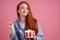 Cute red long foxy haired ginger woman holding packaging cardboard box bucket with french fries and eating chicken