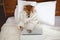 Cute Red Shiba inu dog in the white bathrobe with laptop lying on the bed. Top view