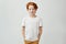 Cute redhead boy with good-looking hairstyle in white t-shirt holding holding hands in pockets, gently smiling and