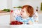 Cute redhead infant baby tasting sweet cherries while sitting in highchair on the kitchen