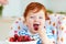 Cute redhead infant baby tasting sweet cherries while sitting in highchair on the kitchen