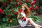 Cute romantic redhead girl with bare feet wearing in a white stylish dress sitting on background of blooming roses and holding a