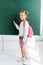 cute schoolgirl standing with backpack and holding chalk near green chalkboard .