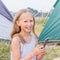 Cute small girl with long blond hair takes a picture on a smartphone and smile on a background of tents