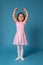 Cute smiling ballerina in a pink dress performs a pose in a ballet dance while standing with raised hands on blue background