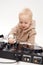 A cute smiling blonde baby in a beige hoodie sits on the floor and plays with dj headphones and a dj mixing board. Music