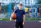 Cute smiling boy in blue t shirt plays basketball on city playground. Active teen enjoying outdoor game with orange ball. Hobby
