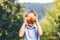 Cute smiling child in blue dress, holding apples in front of her eyes over blurred garden background.