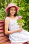 A cute smiling girl in a summer light dress and a pink panama hat sits in the garden on a wooden bench with a rose in her hands.