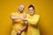 Cute smiling man and woman hug in a friendly way and make a peaceful gesture with their hands. Yellow background.
