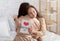 Cute teen girl embracing her mom, congratulating her with Mother`s Day, holding greeting card on bed at home