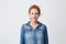 Cute teenage ginger girl with combed hair and beautiful blue eyes standing in denim shirt over gray background