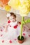 Cute toddler girl in white and red dress sits near huge yellow decorative flower