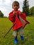 Cute toddler standing on meadow with stick and red farmers jacket