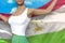 Cute woman in bright skirt holds Tajikistan flag in hands behind her back on the cloudy sky background - flag concept 3d