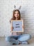 Cute woman in bunny ears holding felt letter board with the words Happy Easter on white brick wall background