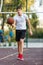 Cute young boy plays basketball on street playground in summer. Teenager in white t-shirt with orange basketball ball outside.