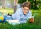 Cute young boy in round glasses and blue shirt lies on the grass, reads book and writes with left hand a homework in the park.