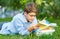 Cute young boy in round glasses and blue shirt lies on the grass, reads book and writes with left hand a homework in the park