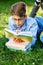 Cute young boy in round glasses and blue shirt lies on the grass, reads book and writes with left hand a homework in the park