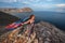 A cute young girl in a dress with a colorful scarf fluttering behind stands on a rock above Lake Baikal.