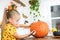 Cute young girl sitting at a table in living room, drawing a face on a large halloween pumpkin. Halloween holiday.