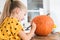 Cute young girl sitting at a table in living room, drawing a face on a large halloween pumpkin. Halloween lifestyle background.