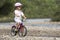 Cute young girl in white clothing, sunglasses with long braids wearing pink safety helmet riding child bicycle on pebbled river