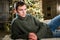 Cute young man in knitted sweater is resting on sofa alone, thinking, a Christmas tree in the background