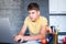 Cute young teenager in yellow shirt sitting behind desk in kitchen next to laptop and study. Serious boy  makes homework,