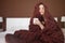 Cute young woman is sitting on the bed wrapped in a big and fluffy brown plaid. Beautiful girl in a cozy blanket