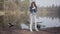 Cute young woman in white pants, jeans jacket and sunglasses standing on the riverbank. The girl is cold, she rubbing