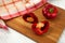 Cutting board with whole and half of red bell pepper and red kitchen towel on white wooden background