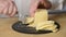 Cutting of cheese into thin slices by means of a cheese slicer on a round chopping board from black slate