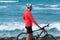 Cyclist wears protective mask, white helmet and red long sleeve stands on ocean shore with mountain bike.