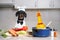 Dachshund dog in chief hat, cooker costume with bow tie sits in kitchen. Tomato, zucchini, carrot and vegetables next to pot with