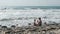 Dad and son is sitting in sea waves in storm on stone beach. Back view.