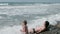 Dad and son is sitting in sea waves in storm on stone beach. Back view.