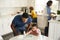 Dad watching his toddler son painting a picture sitting at a table in the kitchen, while mother and girl prepare food in the backg