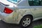 Damaged small gray car parked on a residential street. The rear panel is held on to car with adhesive tape.