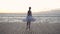 Dancing ballerina in white tutu and pointe on embankment above ocean or sea beach at sunrise. Young beautiful woman